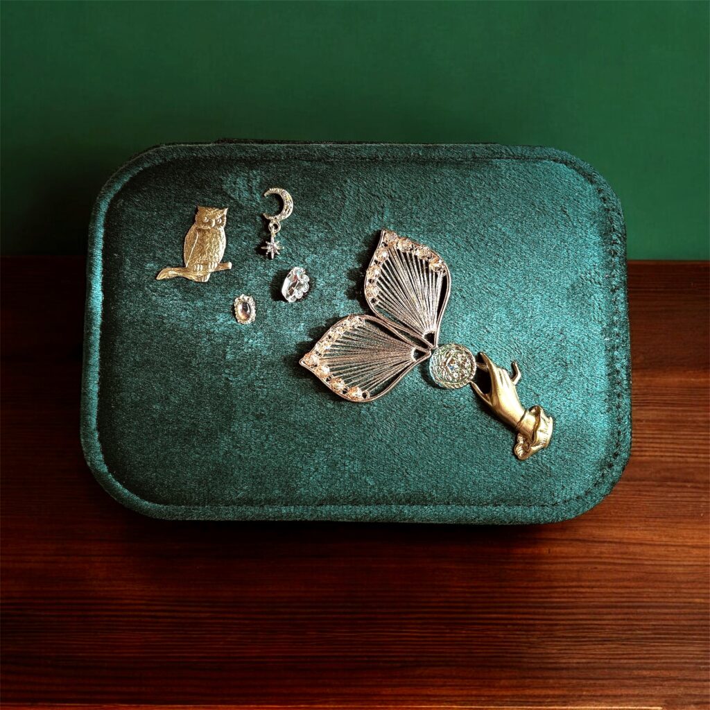 深緑ベルベットのジュエリーケースが木製テーブルの上に置かれ、フェアリーの羽と月・ふくろうのチャームが並ぶFairy Realm Collectionのアクセサリーケース