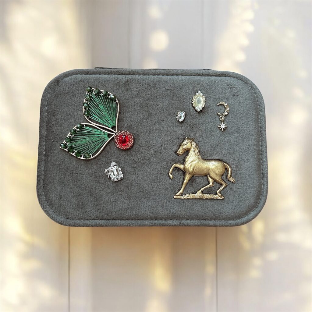 アースグレーのジュエリーケースに、フェアリーを象徴する緑の羽根と赤いガラス、ゴールドの馬、月と星のチャーム、チェコガラスの装飾が並ぶ Fairy Realm Collection の作品
