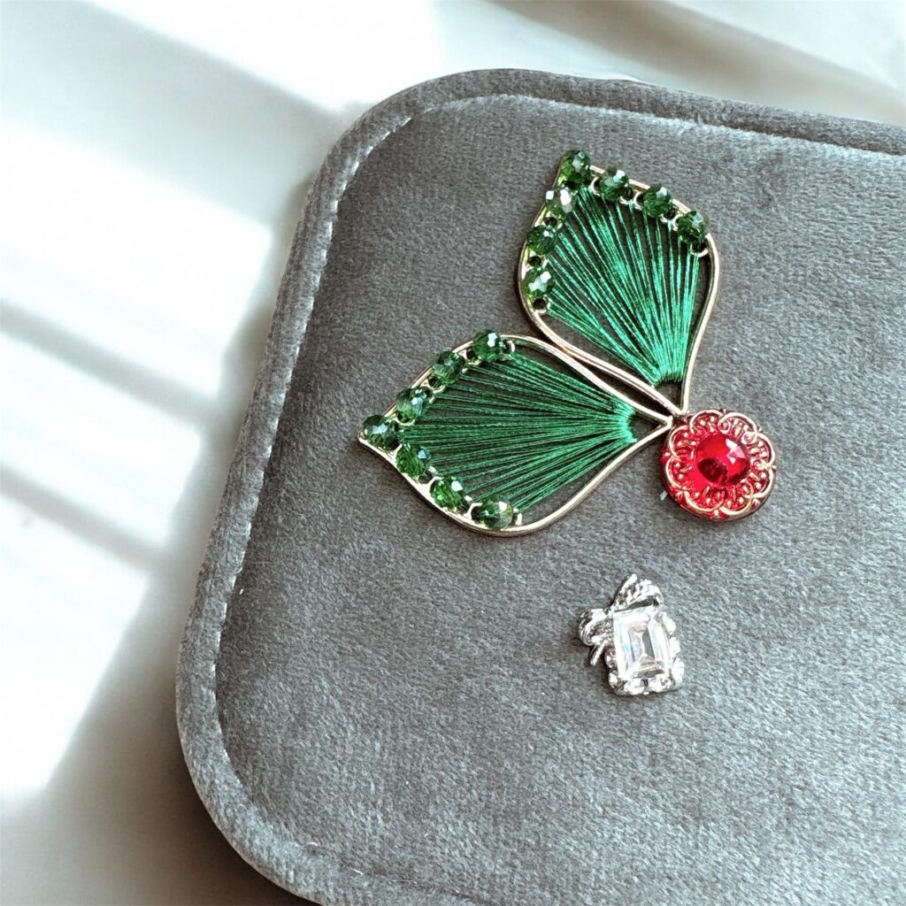 赤いチェコガラスと緑の糸とビーズで形づくられたフェアリーの羽根と、赤いガラスが輝く装飾。アースグレーのジュエリーケースに映える Fairy Realm Collection のデザイン