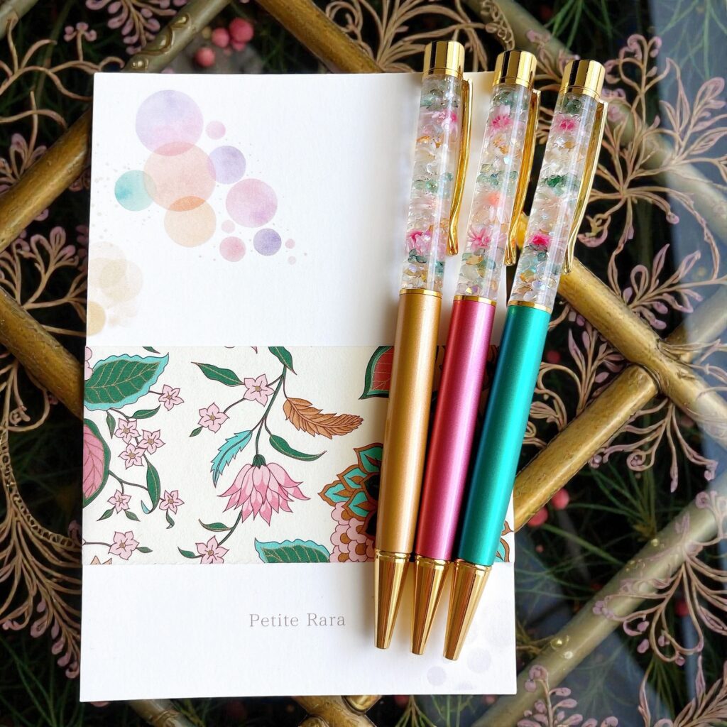 Petite Rara のデザインペーパーの上に、ミント・ゴールド・ピンク軸のハーバリウムボールペンが並ぶ写真。優しい色合いの花材が光を受けて輝く。