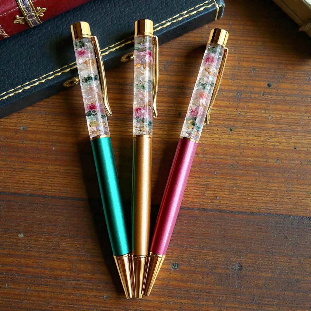 ミント・ゴールド・ピンク軸のハーバリウムボールペンが木製テーブルに並んでいる写真。淡い花材がきらめき、Petite Rara のDewdrop Bloom の雰囲気を表現。