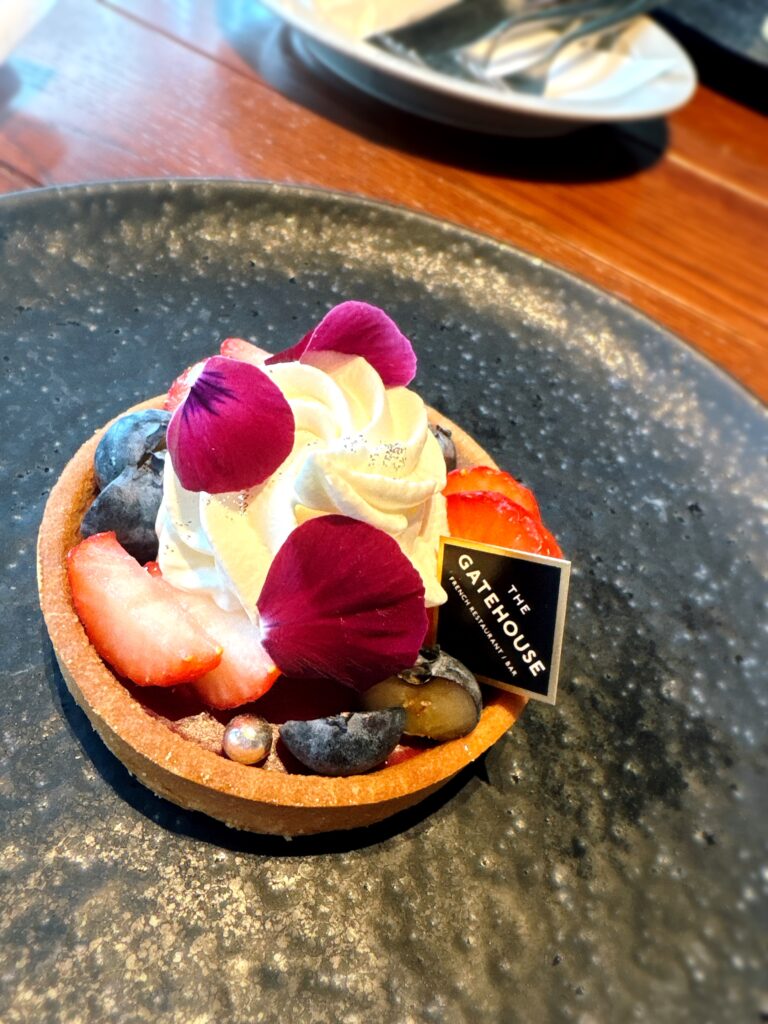 黒い皿にのせられたフルーツタルト。苺やブルーベリー、クリームの上に花びらが添えられ、上品で華やかな印象。
